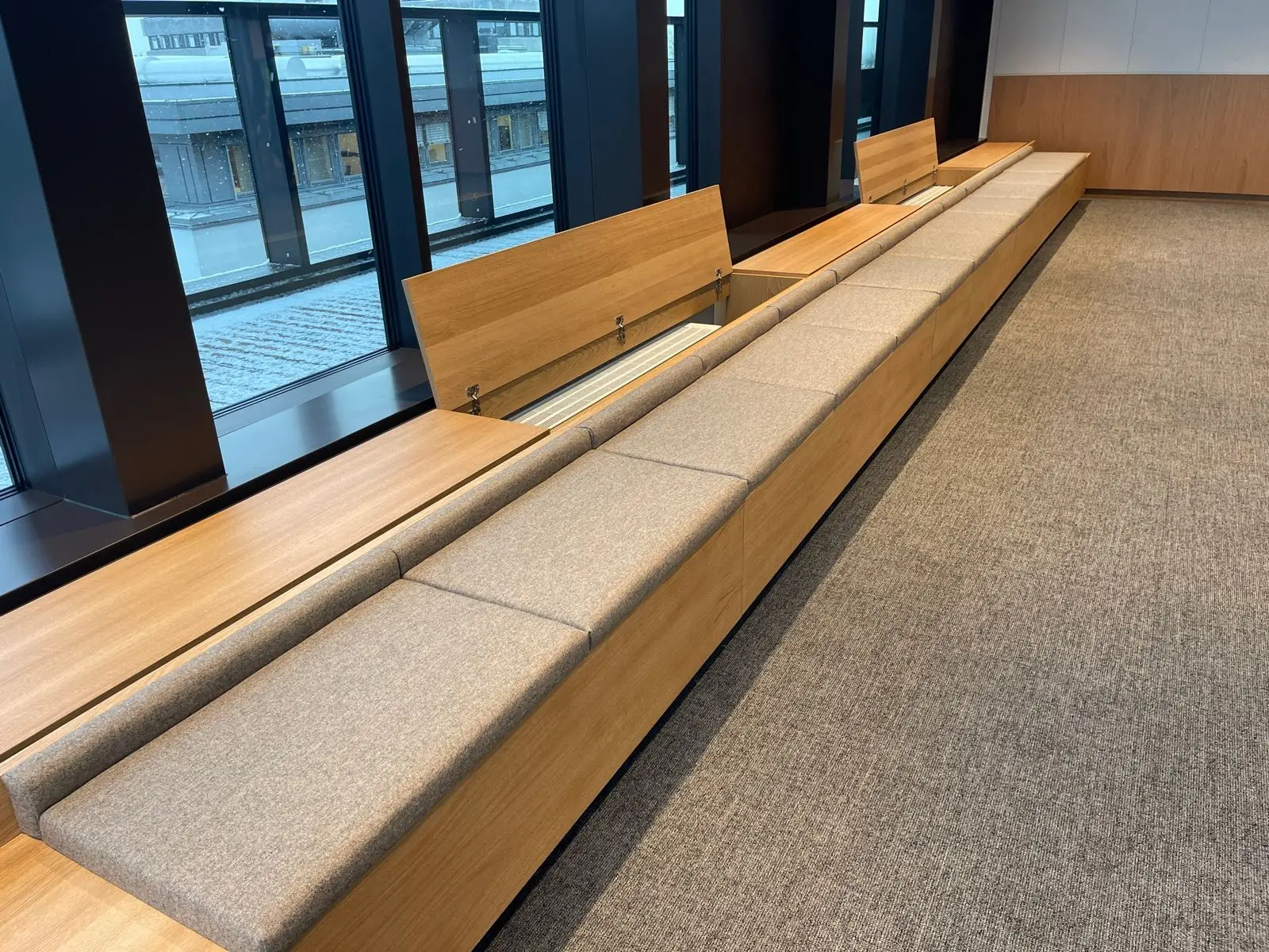 Lang sittebenk med stoppede puter og eikeramme langs vinduene i et offentlig bygg